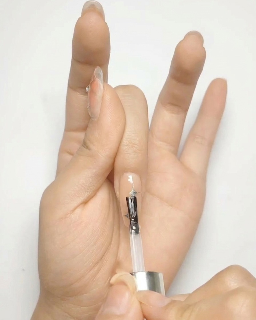 A brush applying a clear, glossy top coat over a nude gel nail decorated with silver stars.