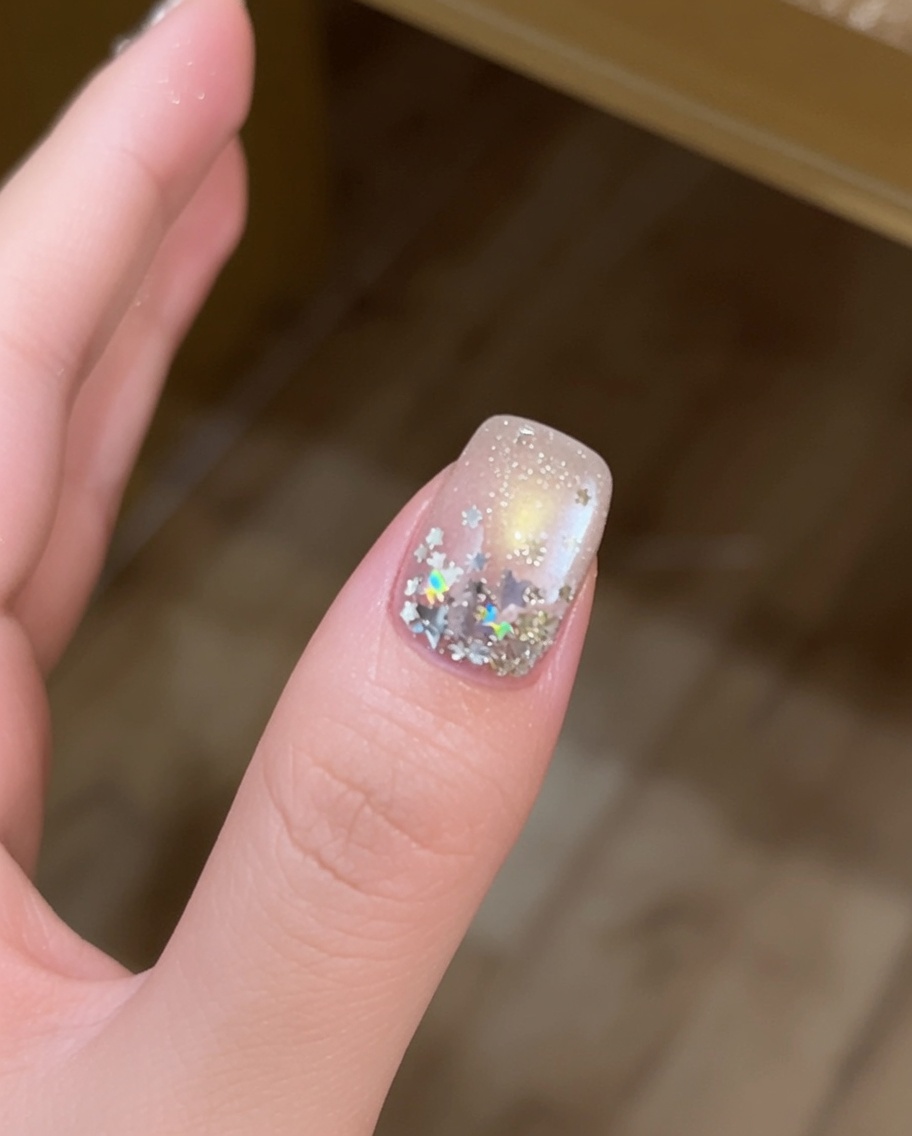 A close-up of a finger with large silver star-shaped sequins being placed onto a gel-coated nail surface with a fine-tipped brush.