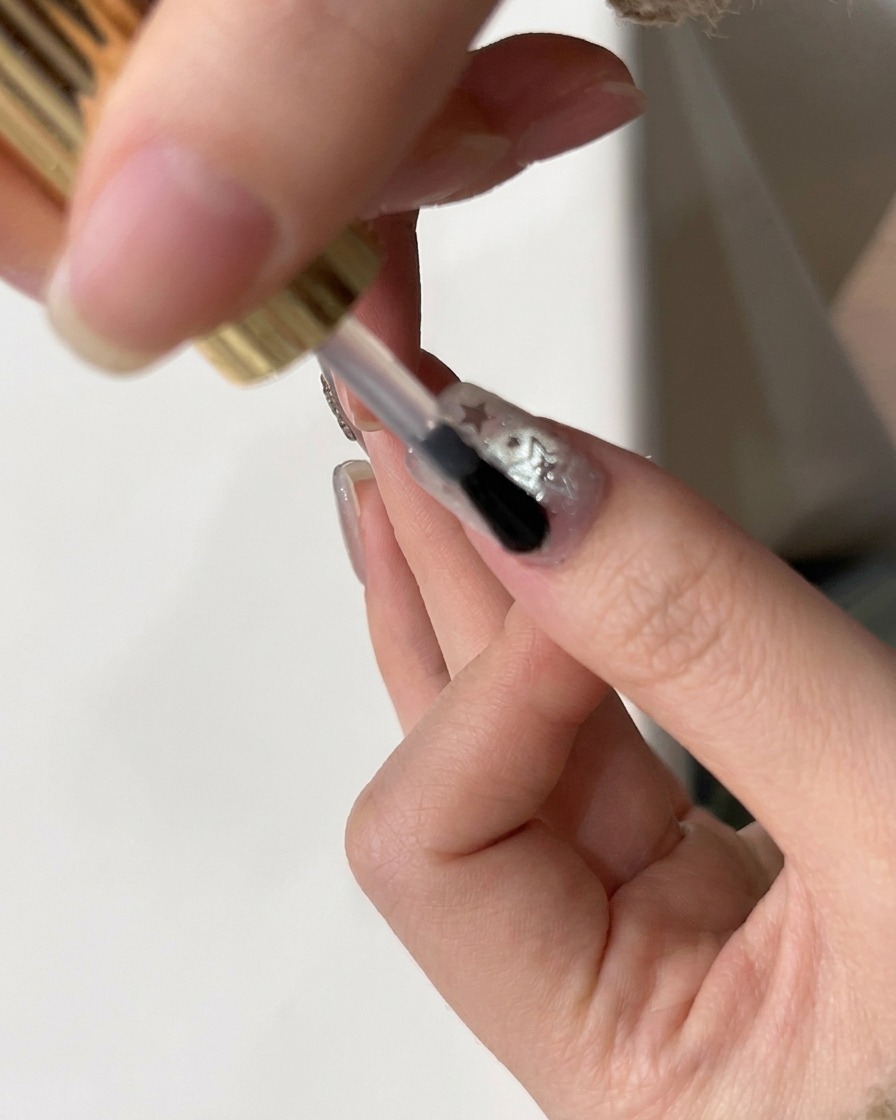 A brush applying a final glossy top coat over a nail decorated with star glitter.
