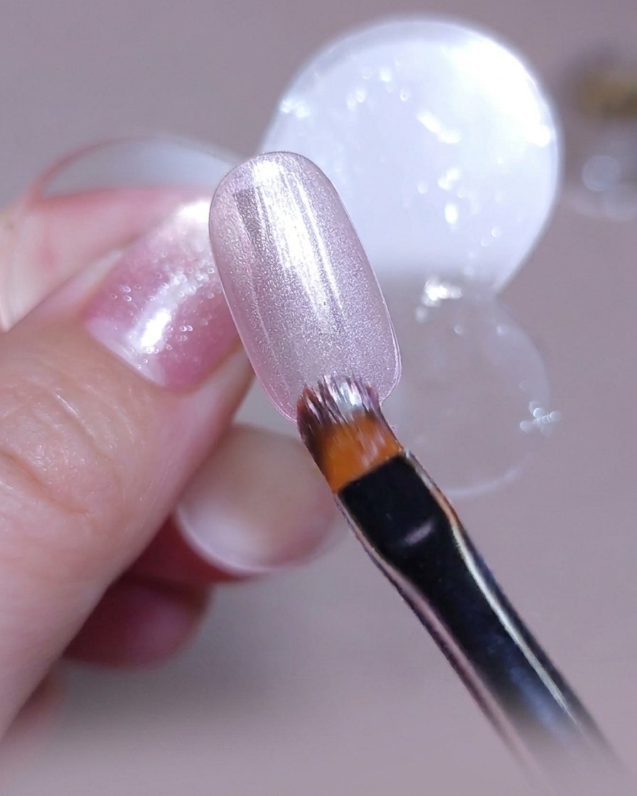 A brush applying a smooth layer of pinkish-silver cat-eye gel polish over a clear nail tip.