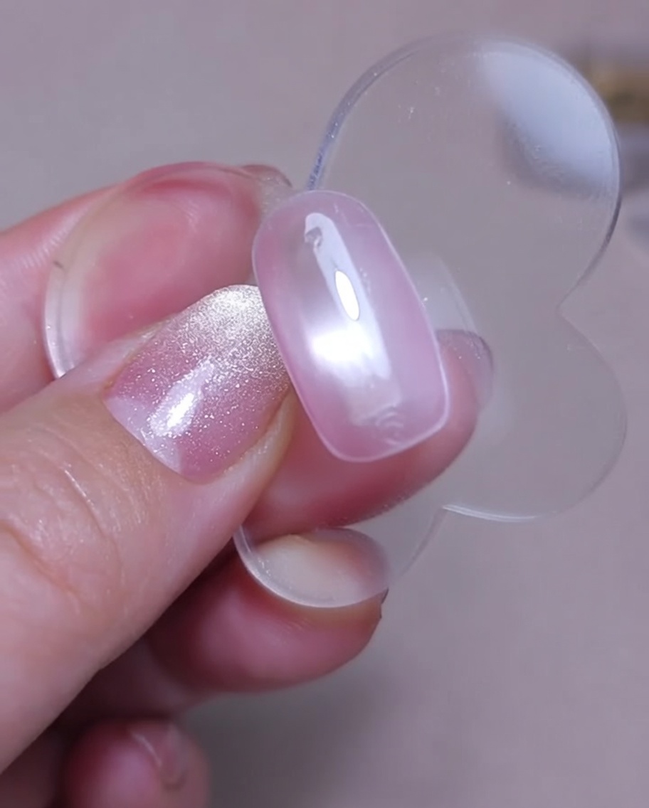 A close-up view of a magnetic nail tool being held over a wet pink cat-eye gel nail to create a shimmering effect.