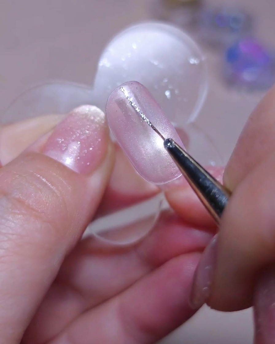 A fine detailer brush drawing a delicate vertical line with silver glitter gel down the left side of a cat-eye nail.