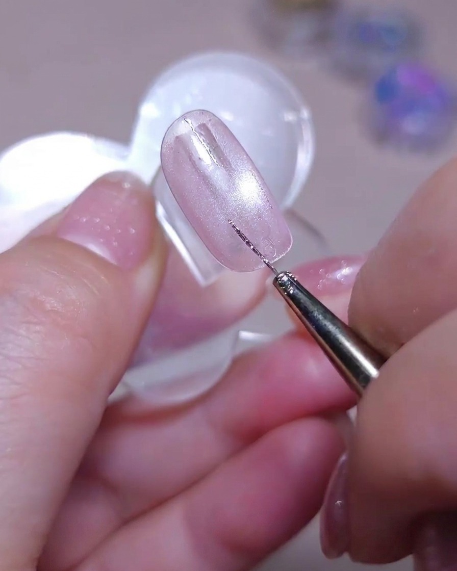 A fine liner brush drawing a second straight vertical silver glitter line next to an existing line on a shimmering pink cat-eye nail.