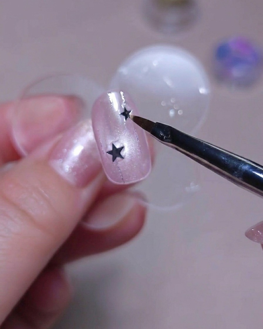 A close-up of a brush tip holding a tiny silver star sequin over a pink glittery nail.