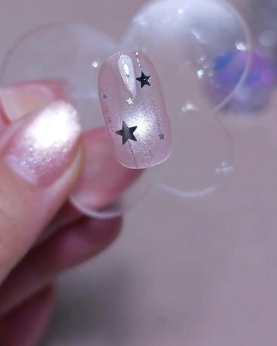 A brush applying a final layer of clear glossy top coat over a nail decorated with vertical lines and silver star sequins.