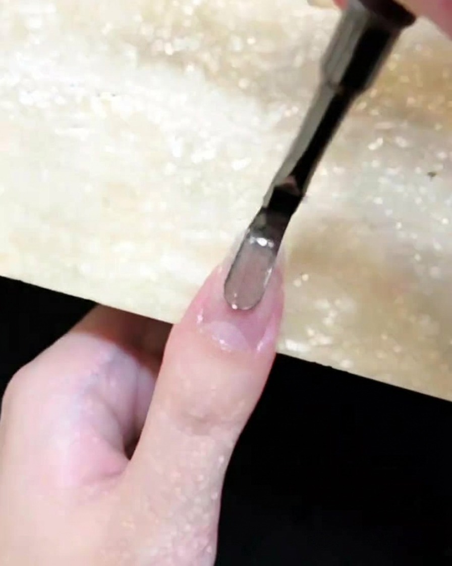 A close-up of a nail brush applying brown cat-eye gel to a natural fingernail.