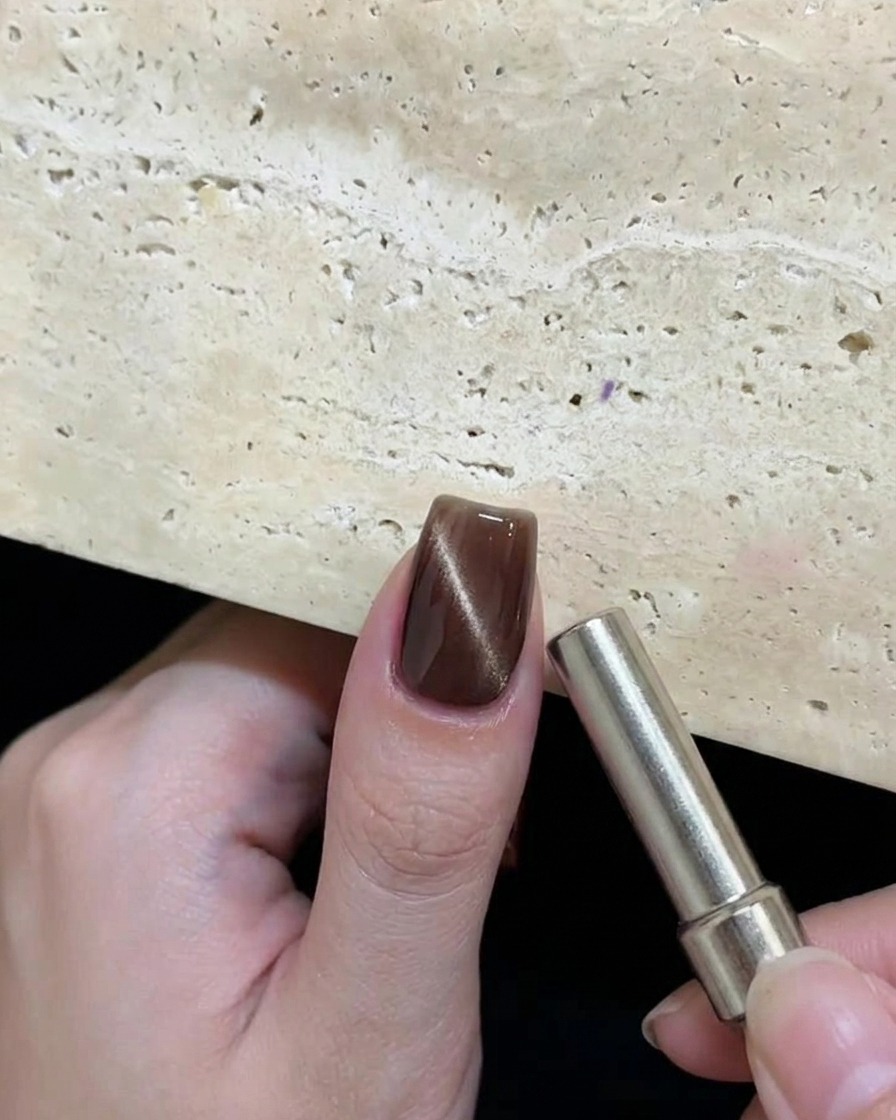 A hand holding a cylindrical magnet near a wet brown cat-eye gel nail to create a circular reflective pattern.