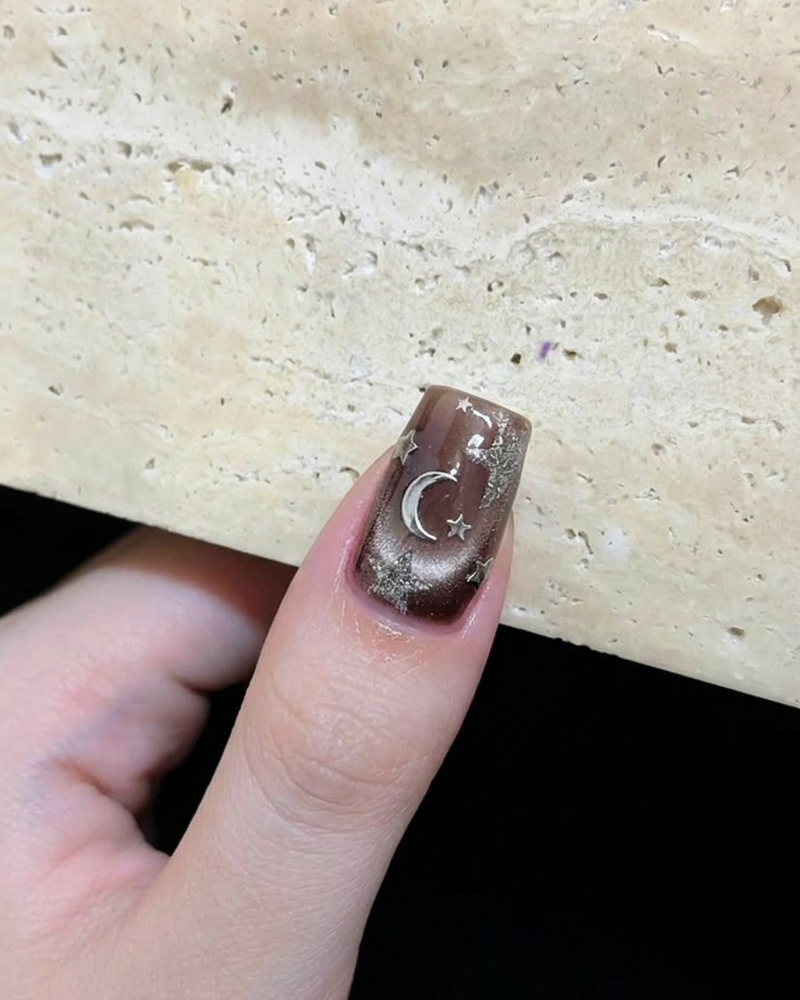 Close-up of a finished dark brown nail with silver stars and a moon accent arranged artistically.