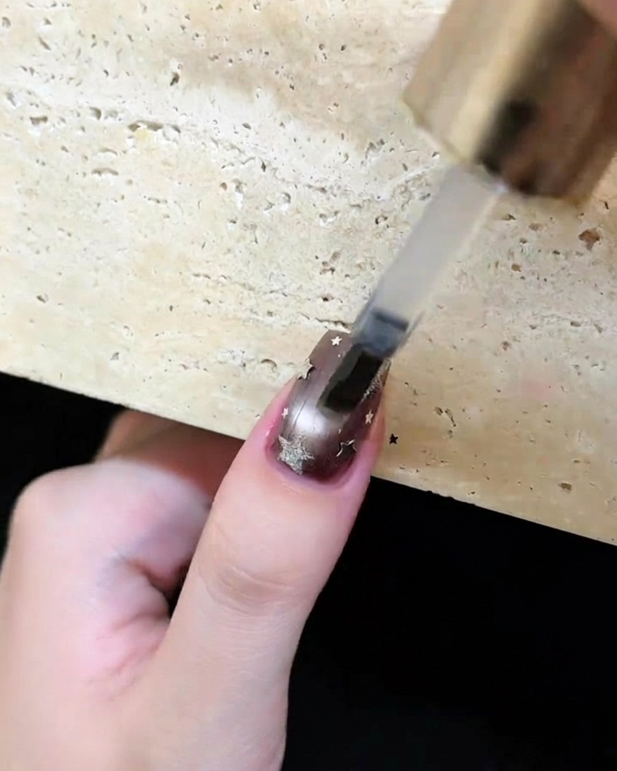 A brush applying a clear, glossy top coat over a dark brown nail decorated with silver stars and moons.