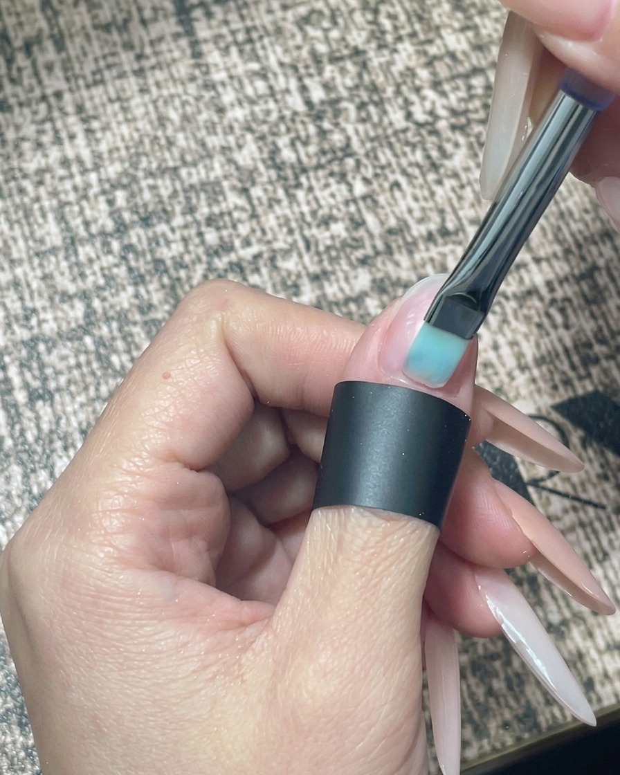 A close-up of a nail brush applying sheer nude gel polish to a prepared, bare natural nail.