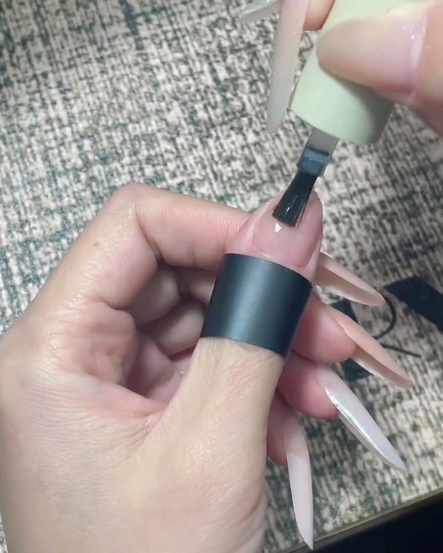 A nail brush applying clear top coat precisely to the tip half of a nude-painted nail.