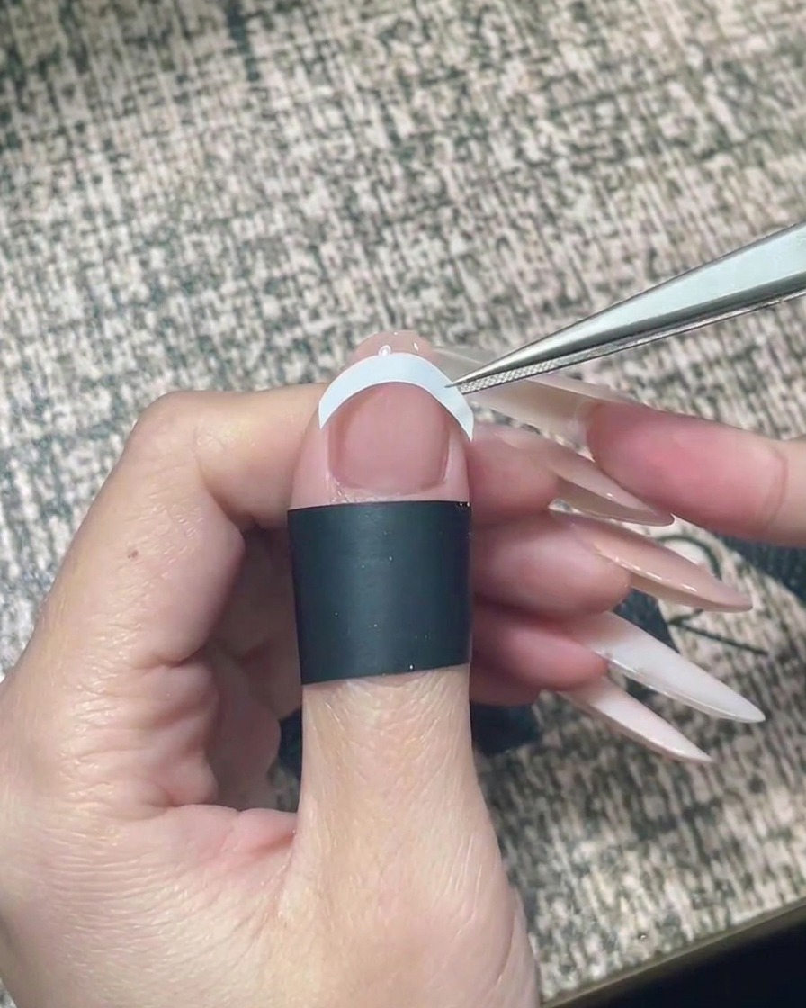 Tweezers placing a white curved French guide sticker horizontally across the mid-section of a nail.