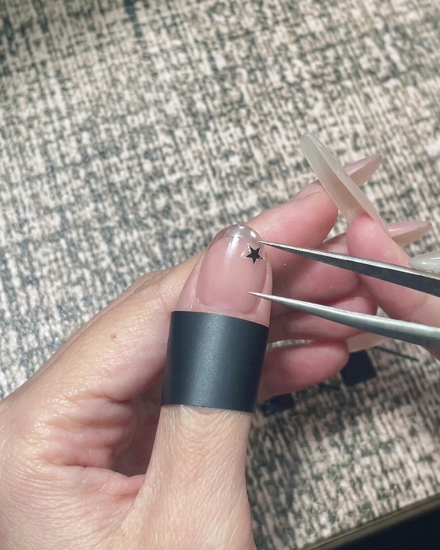 A hand holding a pair of tweezers to place a small black star sticker onto a nude gel nail.