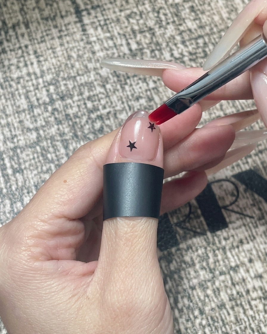 A square gel brush applying clear encapsulating gel over a nail decorated with black stars and a chrome tip.