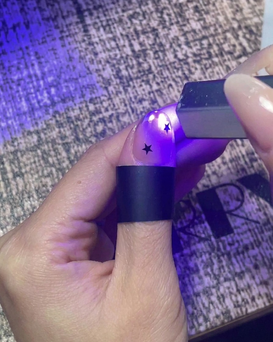 A close-up view of a hand using a small handheld UV flashlight to flash cure a nail decorated with black star stickers.