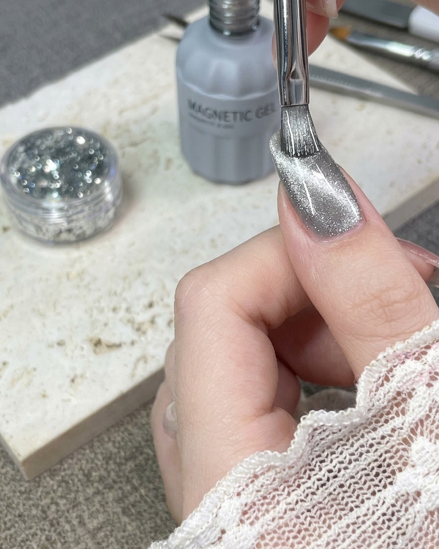 A brush applying a layer of silver cat-eye gel polish onto a natural fingernail.