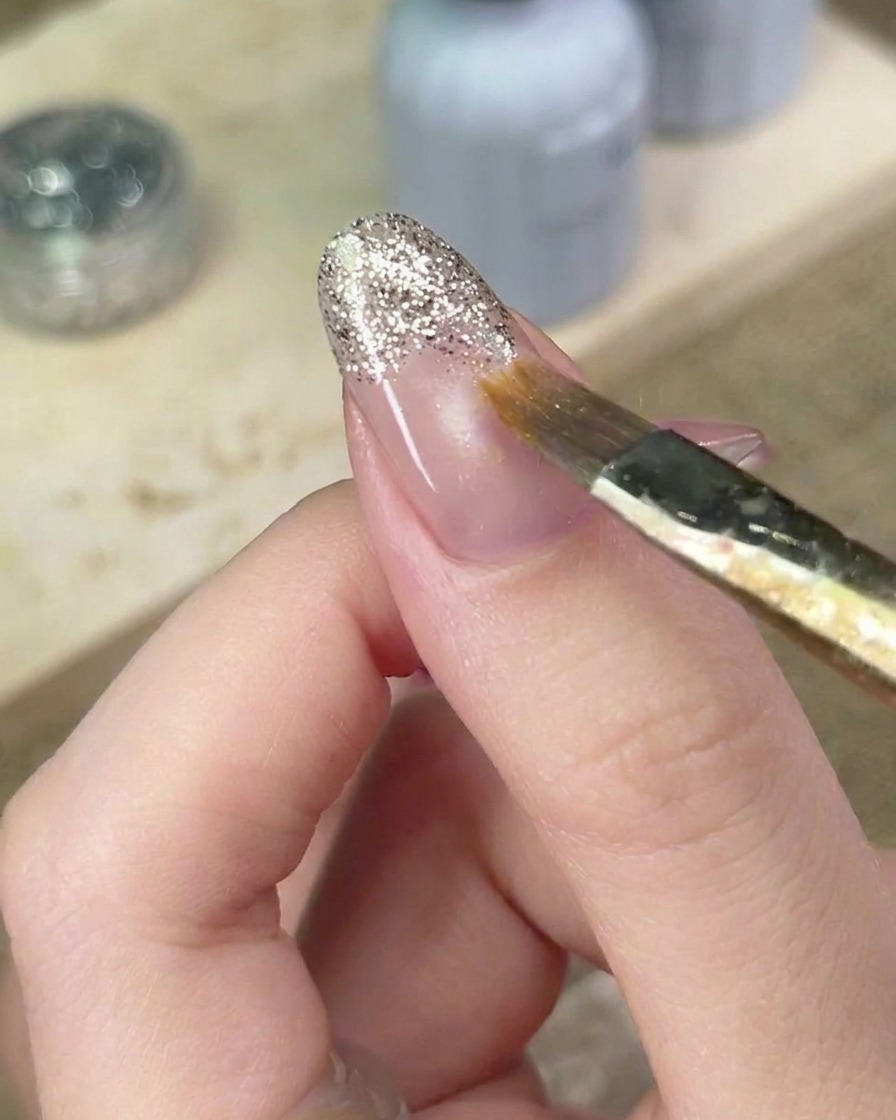 A detail brush carefully blending the edges of silver glitter gel on the nail to perfect the gradient.
