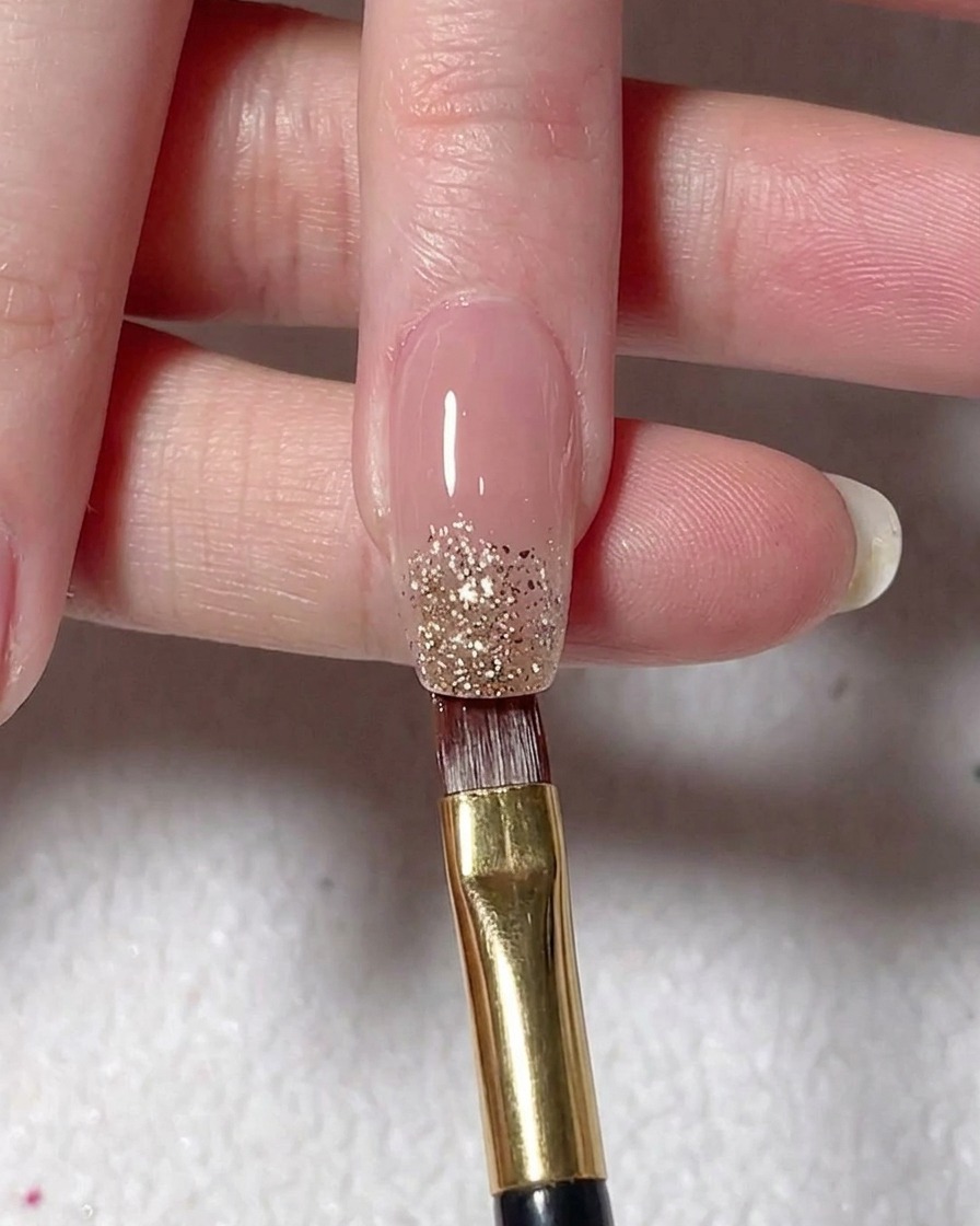 A flat brush depositing silver glitter gel onto the tip of a nude-painted nail.
