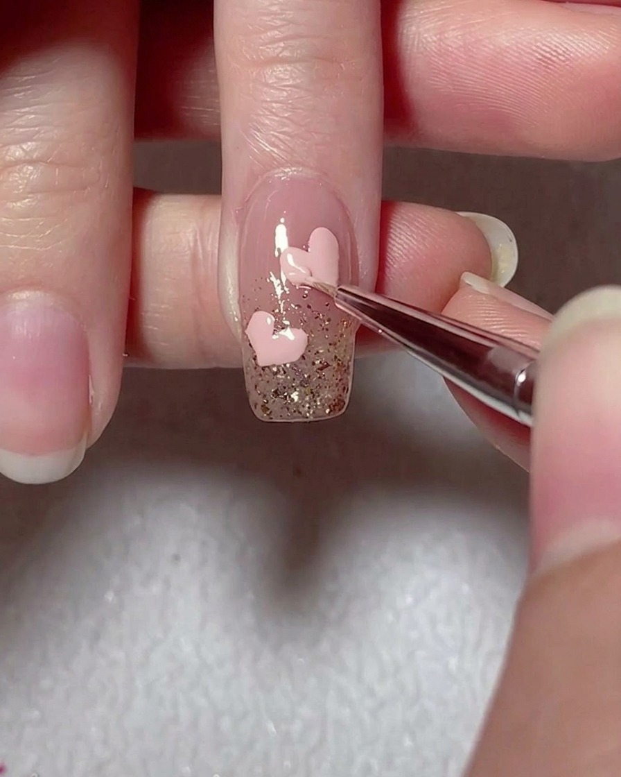 A liner brush refining the edges of a pink gel heart on a glitter-accented nail.