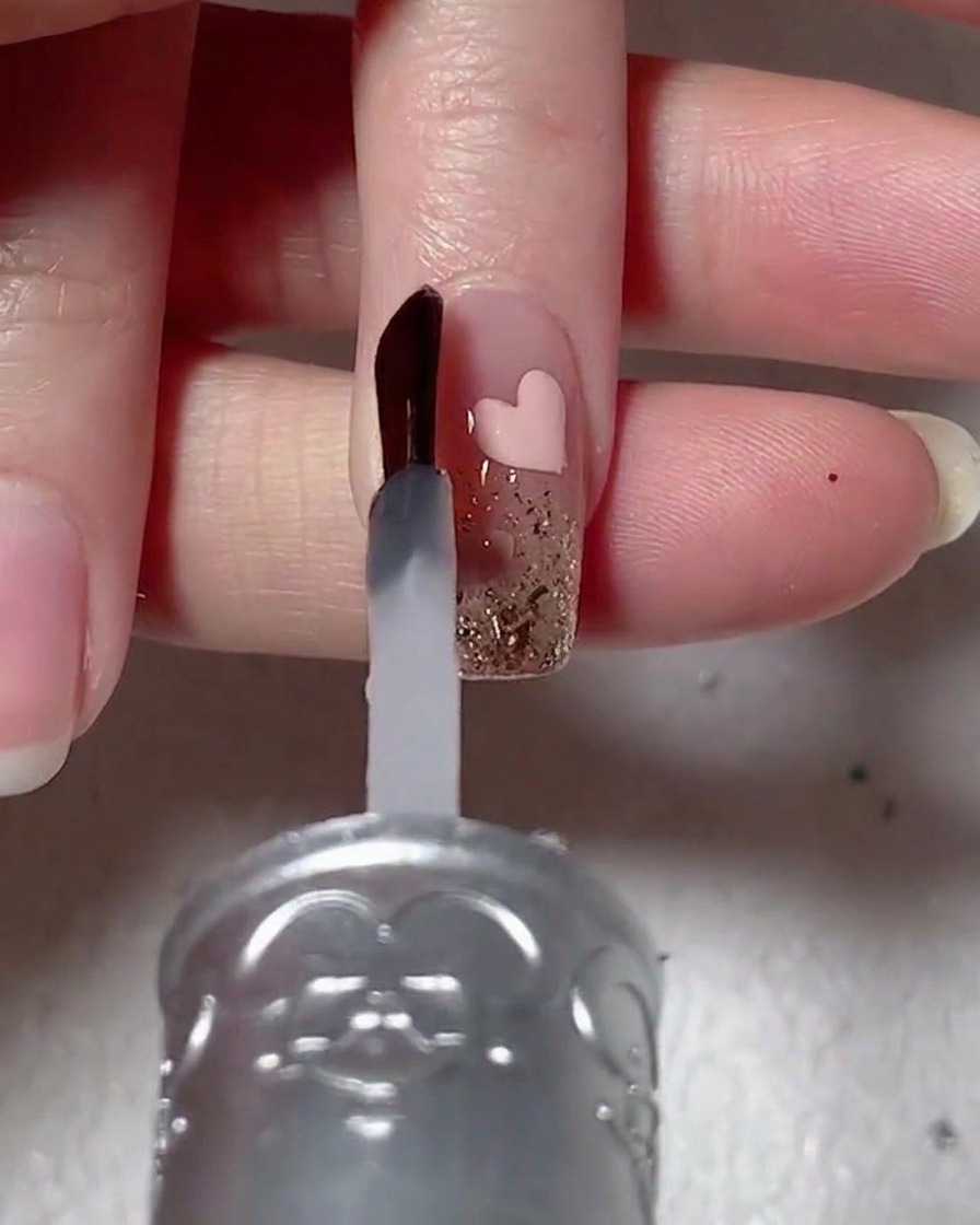 A nail brush applying a clear, viscous reinforcement gel over a nail decorated with glitter and a pink heart design.