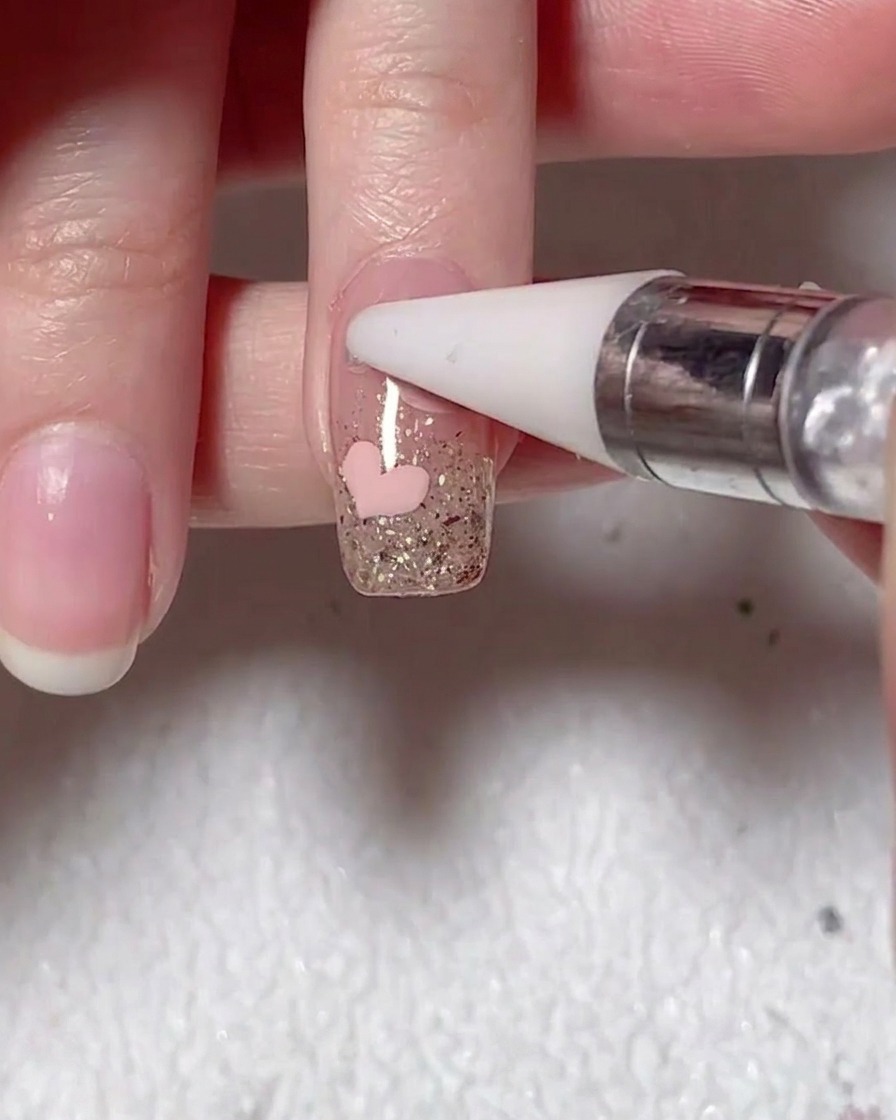 A wax-tipped tool placing a small pearl onto the surface of a nail next to a painted pink heart.