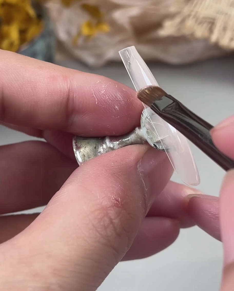 A nail brush applying a milky nude gel base coat to a clear nail tip held by fingers.