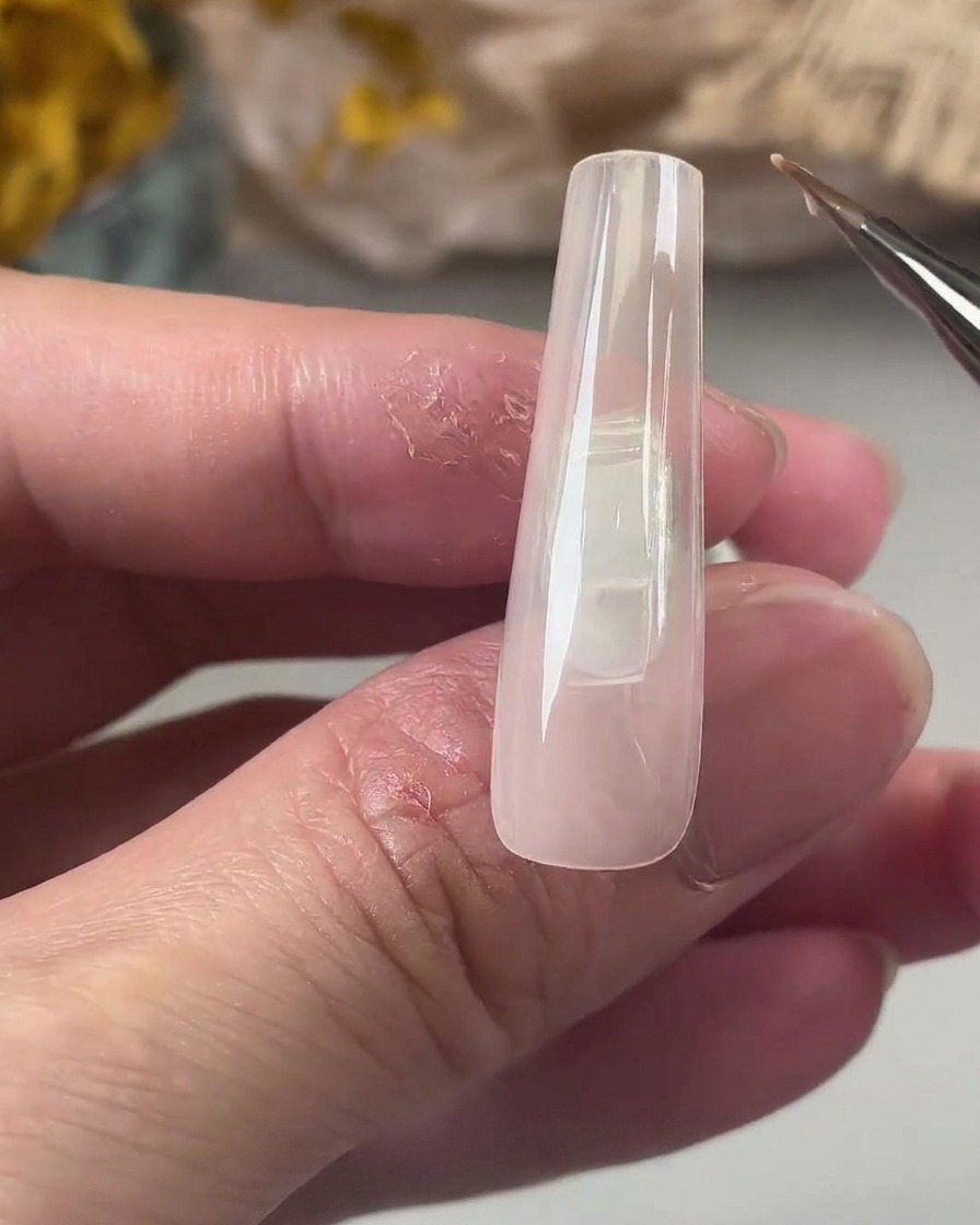 A fine detail brush applying a smooth layer of clear base coat gel onto a long, nude-colored square nail tip.