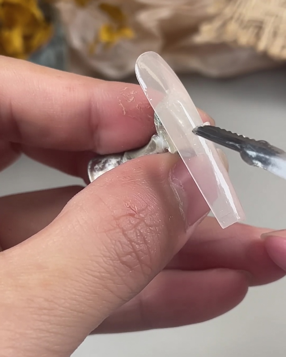 A hand using a tool to carefully place iridescent, holographic glitter flakes onto the wet, uncured gel of a nail tip.