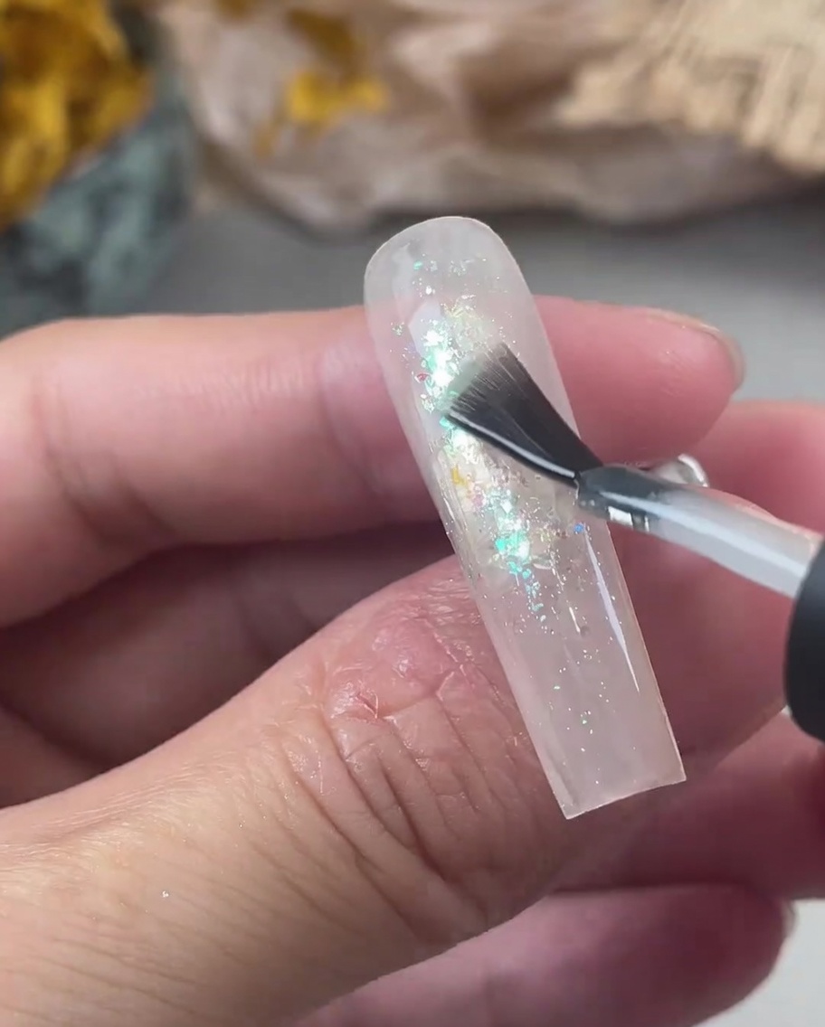 A nail brush applying a thick layer of clear strengthening gel over an iridescent glitter nail.