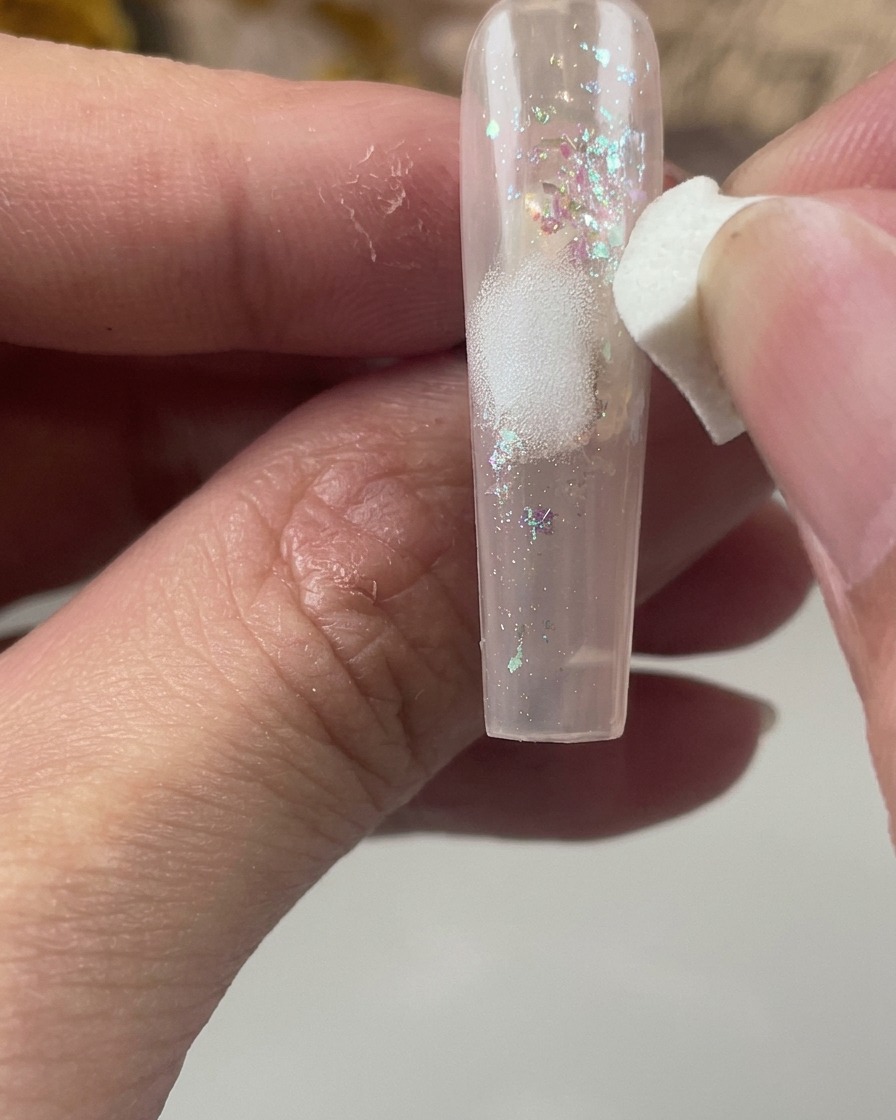 A white cosmetic sponge gently dabbing a soft patch of white paint onto the center of a glitter-encapsulated nail.