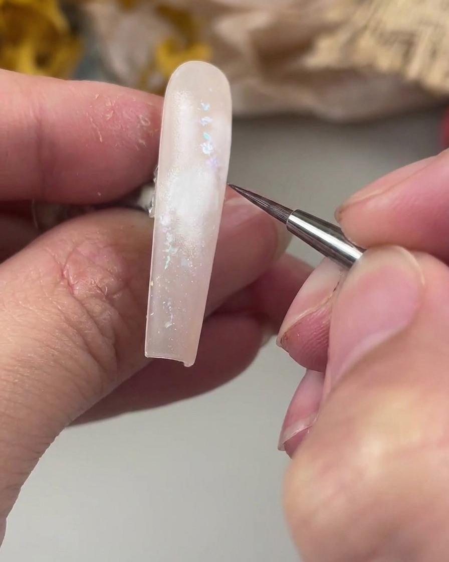 A fine liner brush poised over the white painted area on the nail, ready to paint the delicate watercolor rose center.