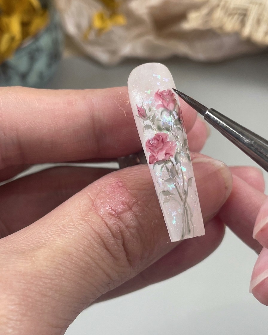 A close-up view of a fine nail art brush painting a small, soft pink water-drop shape onto a nail surface as part of a floral design.