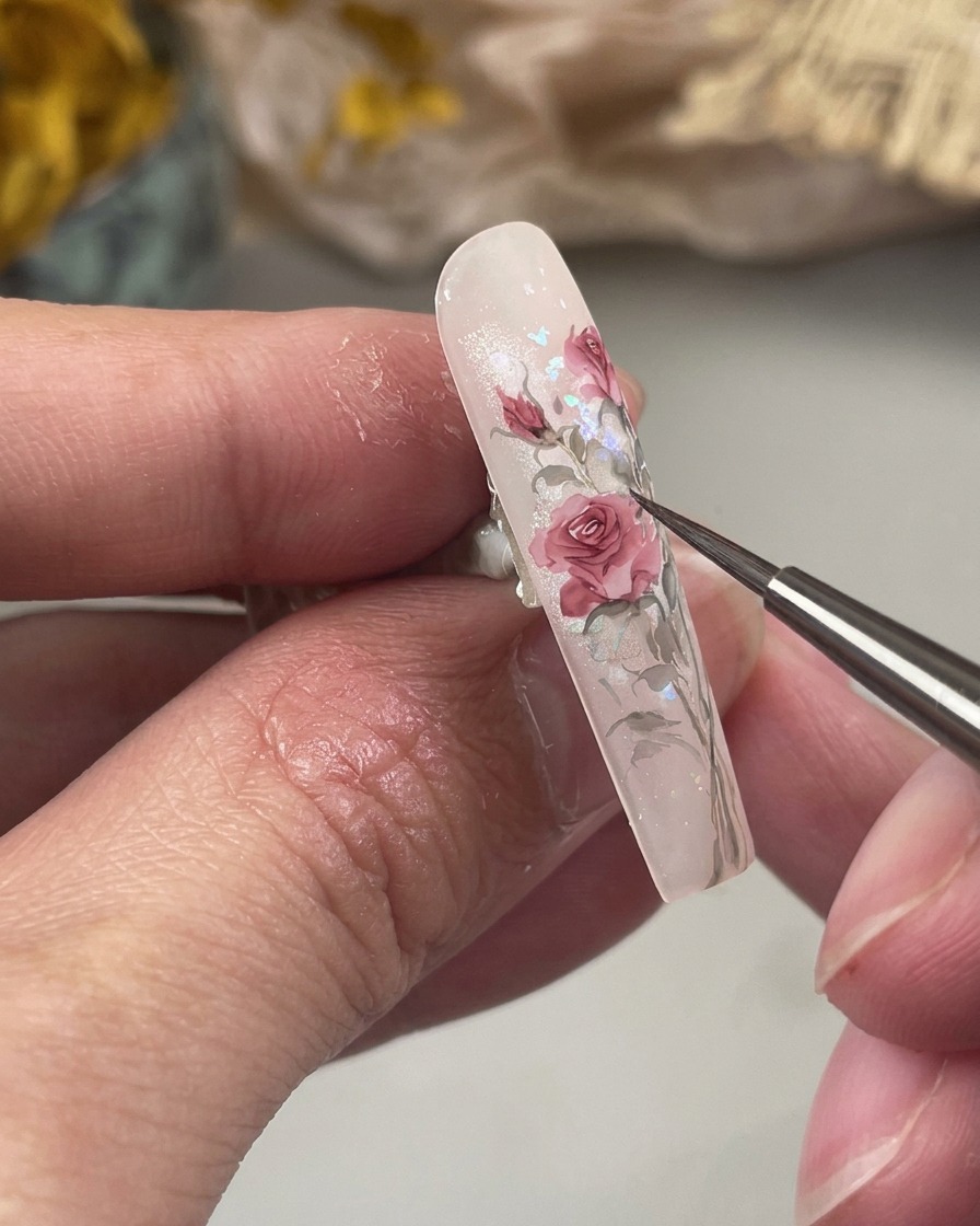 A close-up shot showing a fine brush painting small, tapered green leaves at the base of rose-colored floral art on the nail.