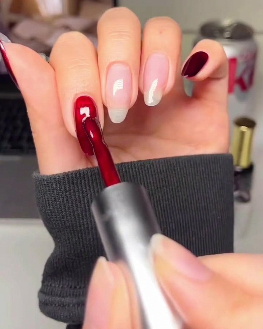 A close-up of a nail brush applying a deep, rich red gel polish to a prepared nail surface.