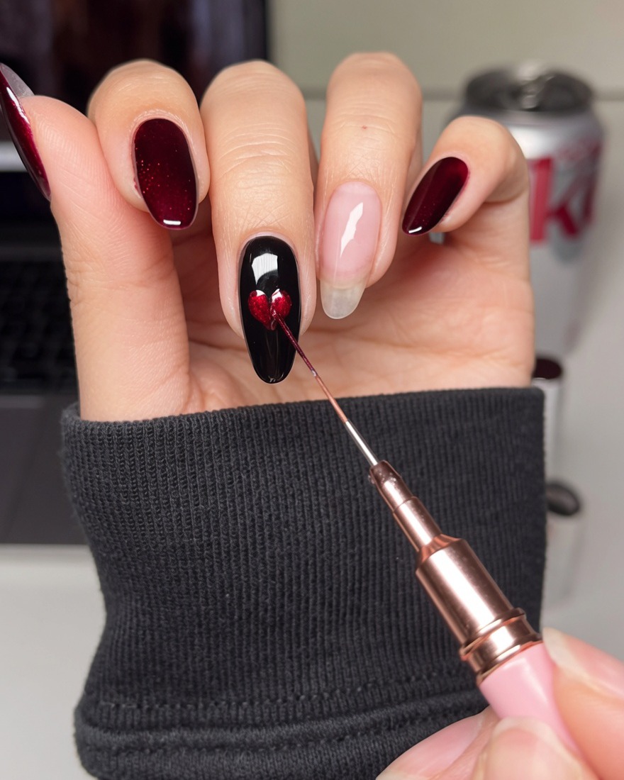 A fine detail brush filling in the center of a heart-shaped stencil on a black nail with red gel polish.