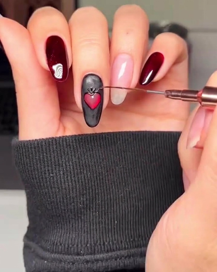 A close-up view of a black nail featuring a red heart, with a fine brush adding gold decorative accents around the hearts perimeter.
