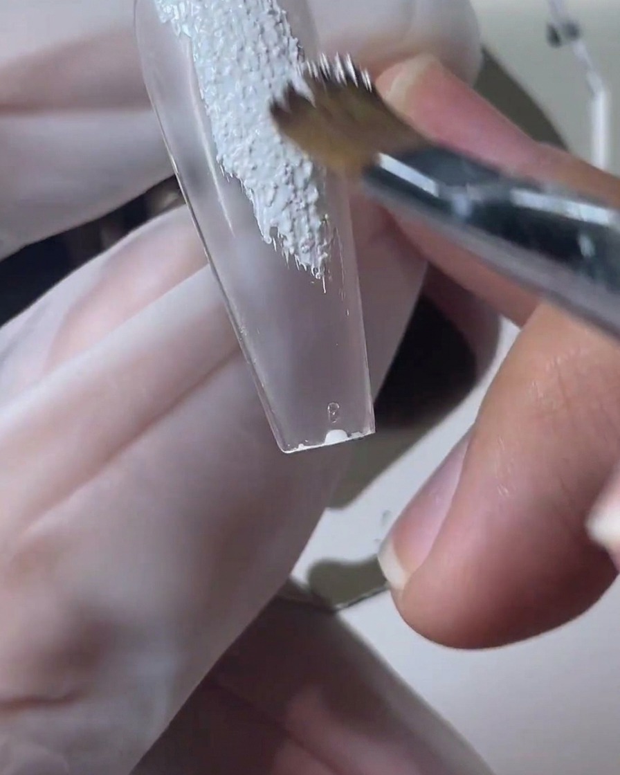 A nail brush applying textured white sand gel over a clear nail tip.