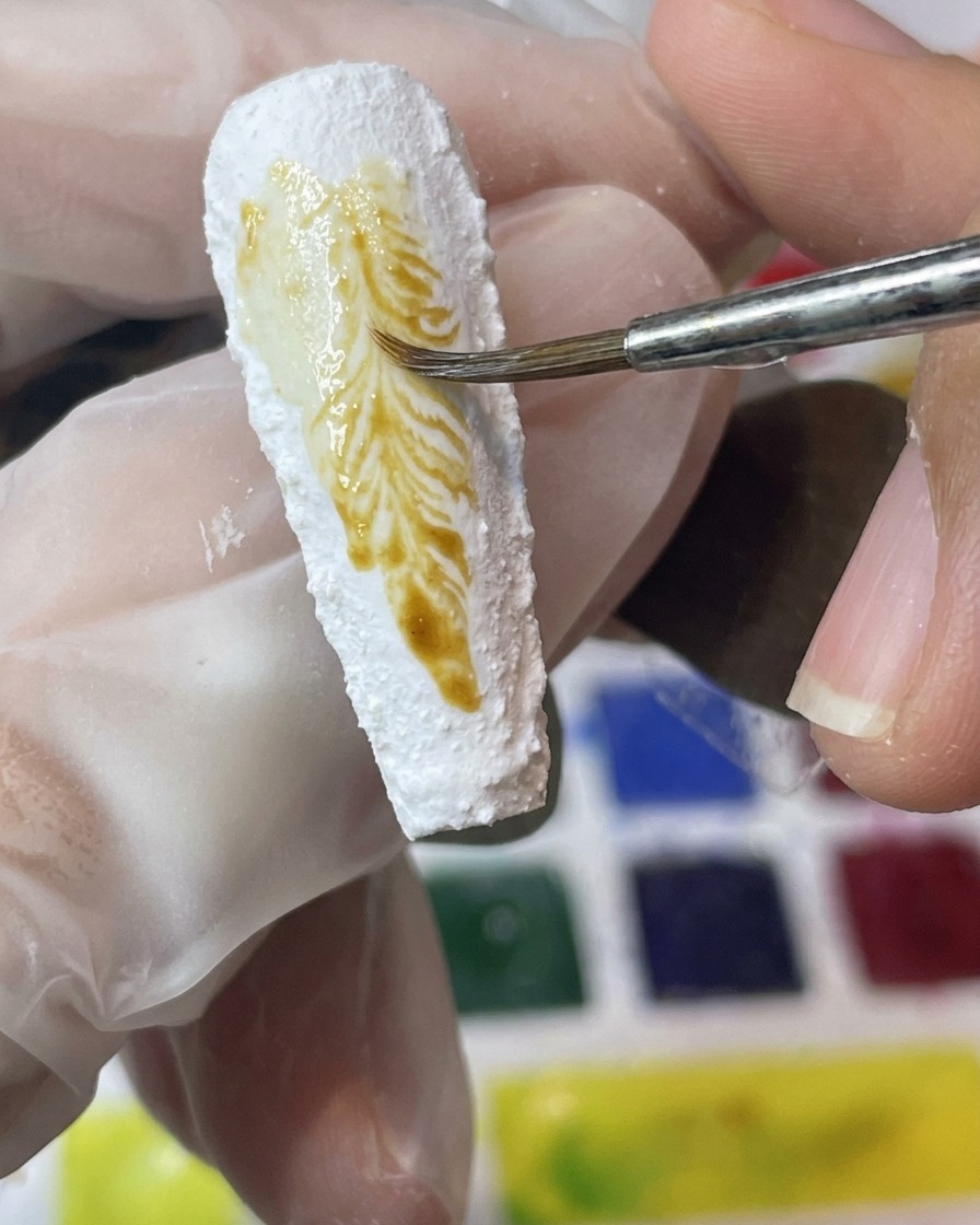 A brush applying yellow and brown pigments to a textured white nail surface.