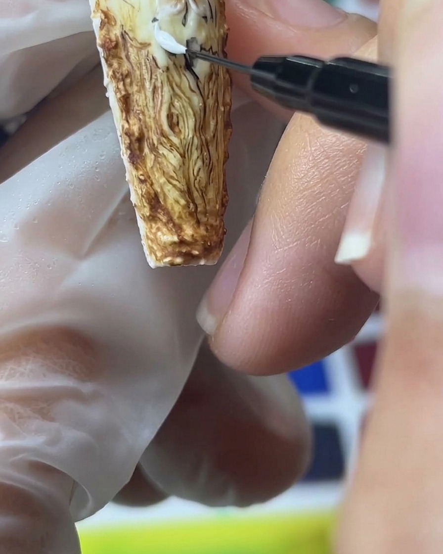 A close-up view of a fine detail brush applying white sand gel to create textured, dimensional rose petals on a brown background.