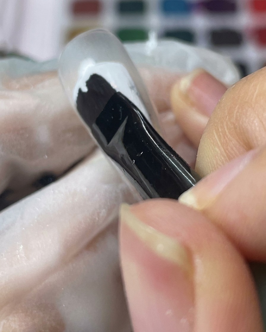 A black gel brush applying a smooth coat of white gel to a clear nail tip.