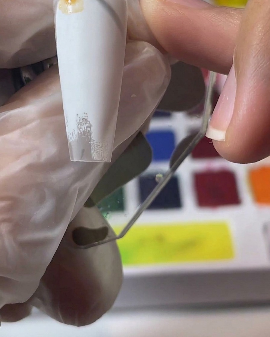 A hand holding a white nail tip while mixing and applying brown watercolor pigments.
