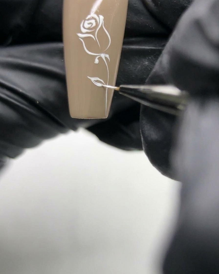 Macro shot of a nail artist refining the delicate white lines of a rose design on a neutral base nail.