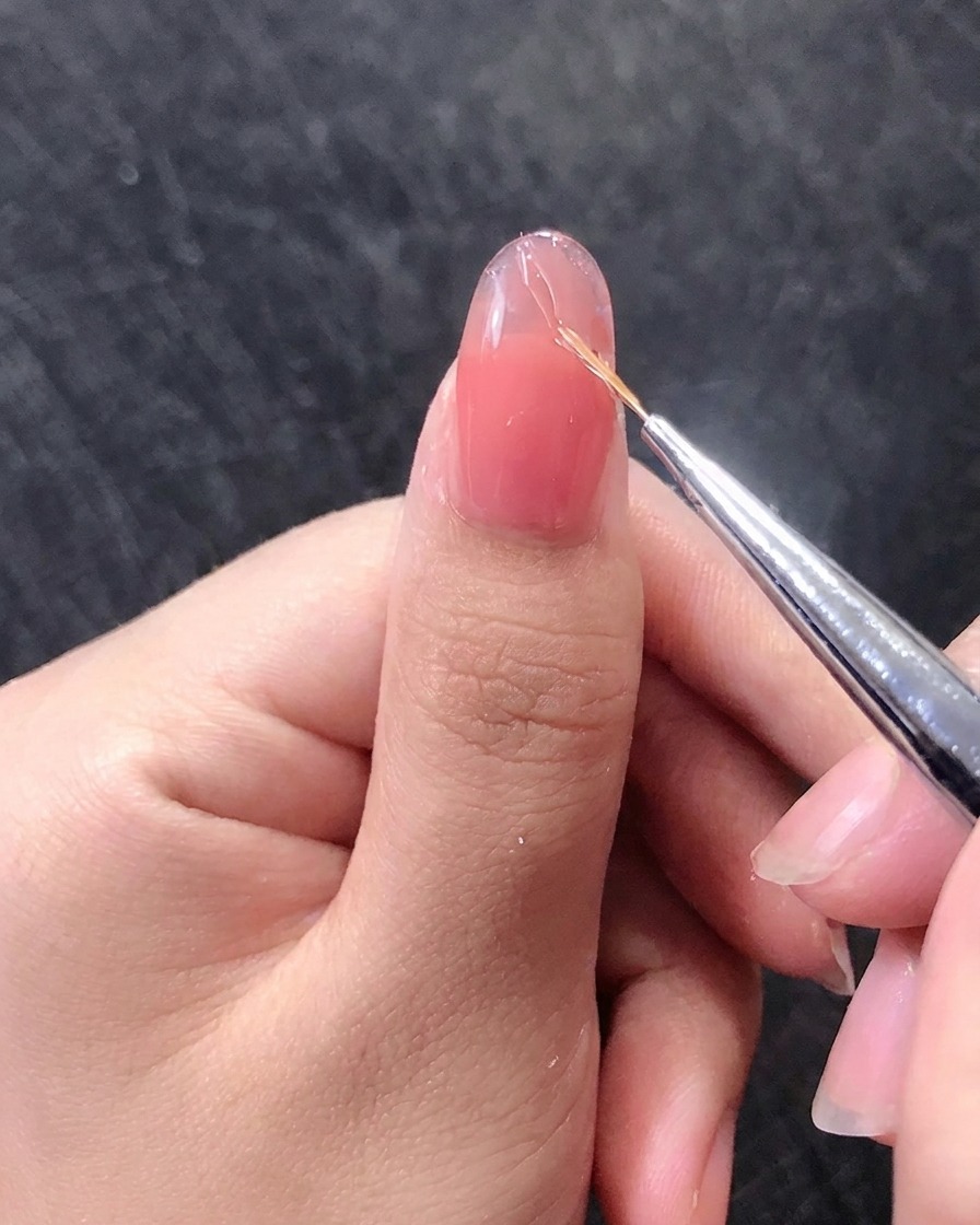 A close-up of a nail brush drawing fine gold lines on a pink gel nail.