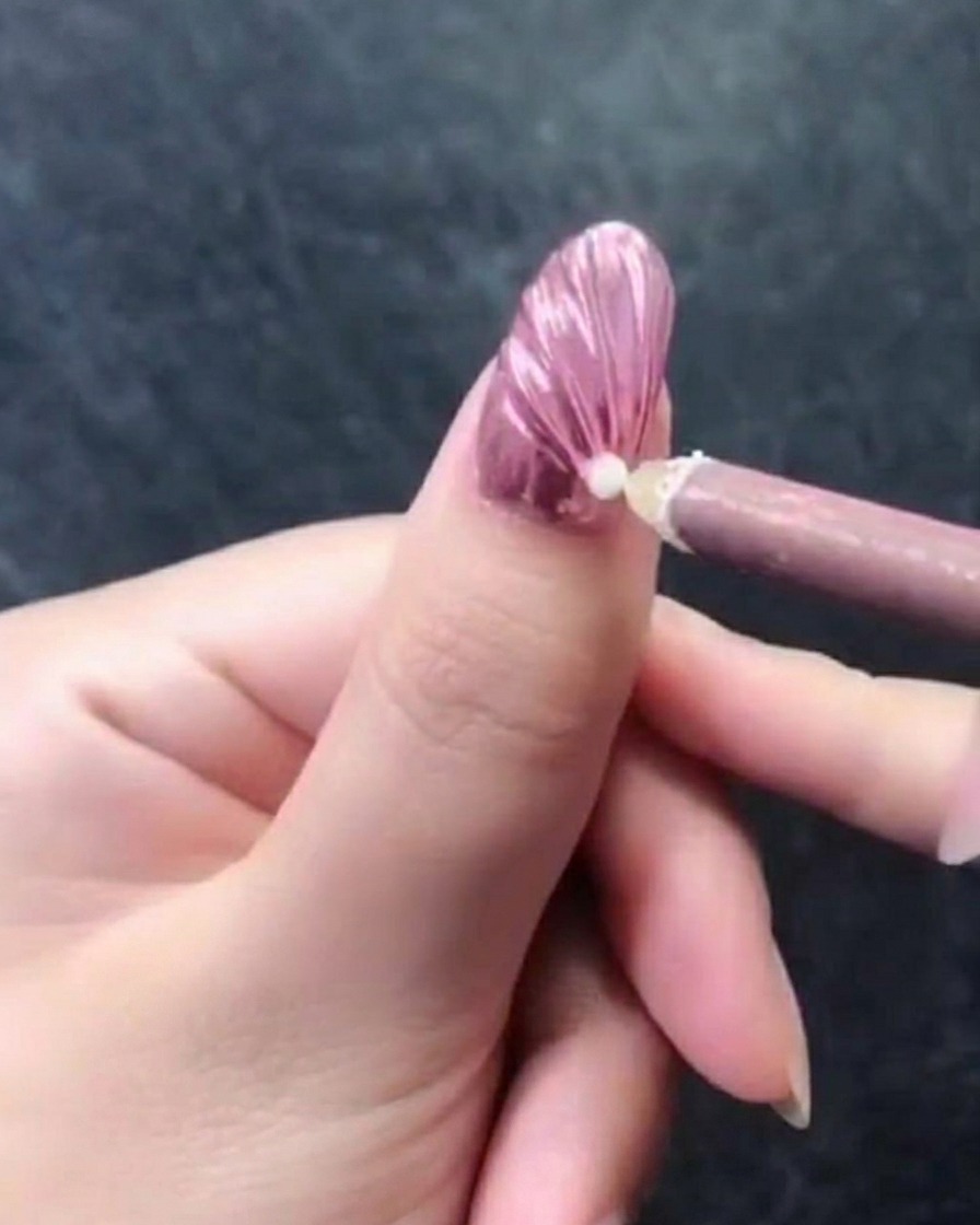 A close-up of a hand with a pink metallic textured nail, showing a pink wax picker pen placing a single white pearl near the cuticle area.