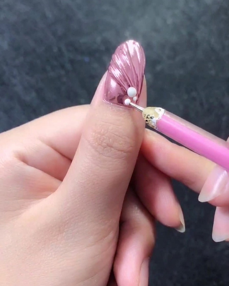 A close-up of a pink metallic textured nail with a wax picker pen adding tiny white beads directly next to a larger white pearl at the cuticle.
