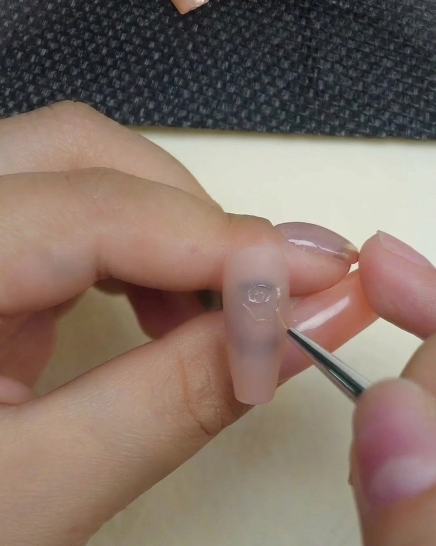 A close-up of a hand using a fine liner brush to apply clear 3D sculpting gel, drawing the precise center swirl of a rose onto a matte nude nail.
