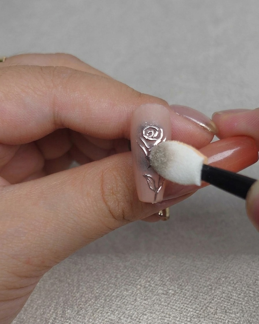 A close-up of a sponge buffing chrome powder into the grooves of a 3D rose nail design, creating a high-shine metallic finish.