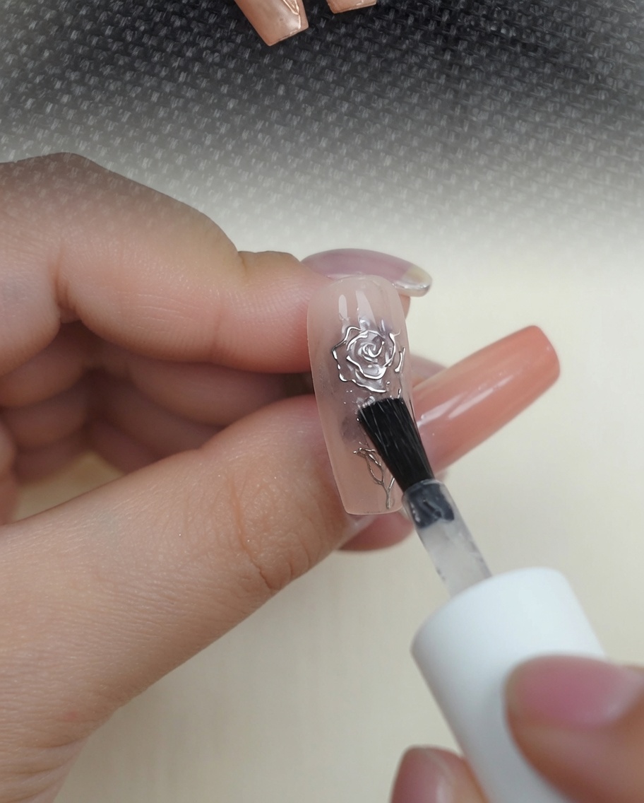 A brush applying a clear, glossy top coat over a metallic chrome 3D rose design on a nude nail.