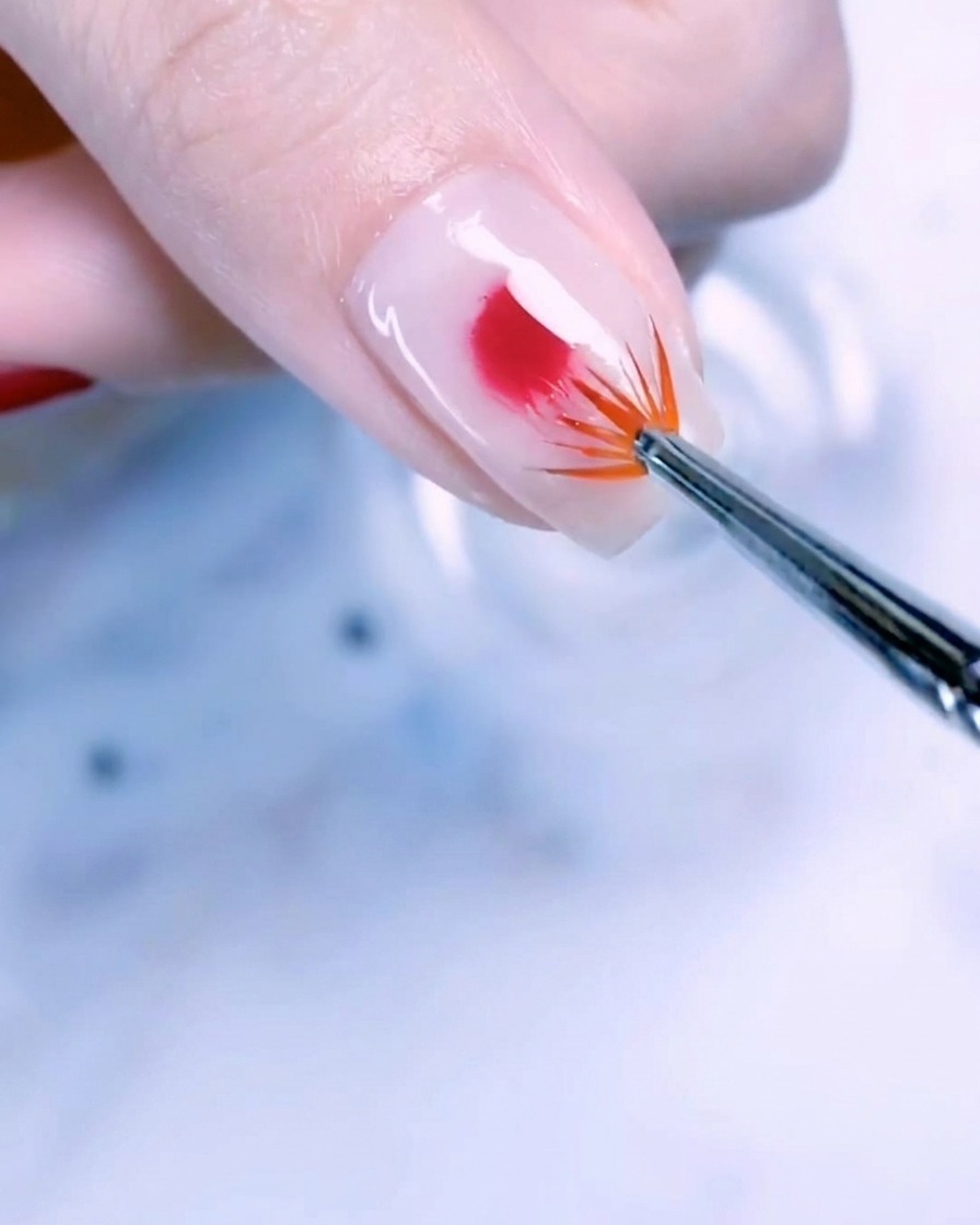 A close-up of a thumbnail with a milky white base and a centered drop of rose gold gel polish being blended outward with a fan brush.