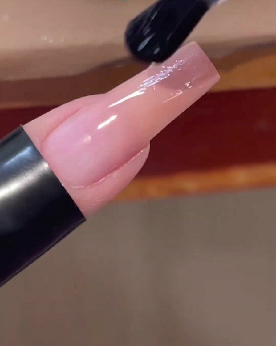 A nail brush applying a clear, glossy top coat over the finished pink gradient nail.